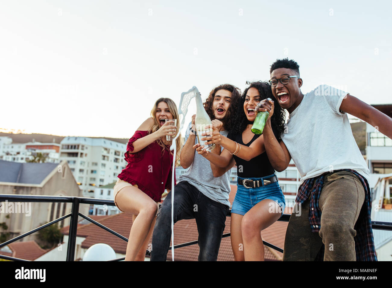 Cheerful friends celebrating rooftop party with champagne. Women opening the champagne bottle with her friends screaming and enjoying. Young men and w Stock Photo