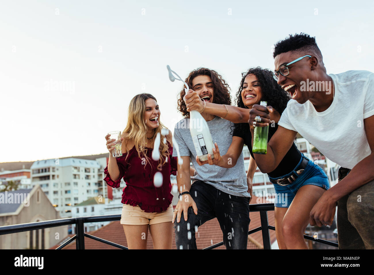 Outdoor shot of woman opening a bottle of champagne with friends enjoying. Group of friends having champagne at rooftop party. Stock Photo