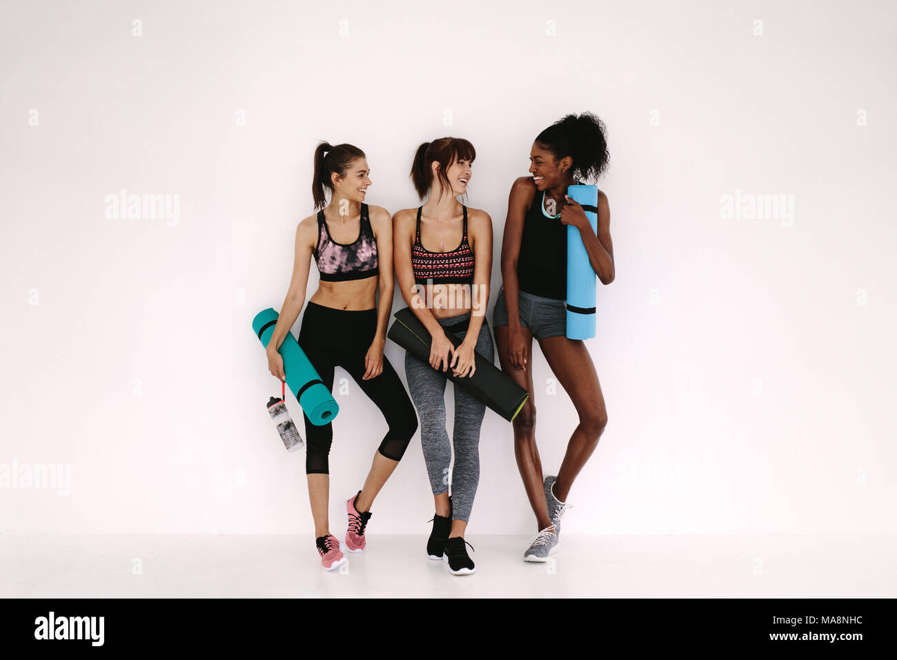 Group of female friends in sportswear smiling together while standing in a gym after yoga workout. Women standing by a wall with exercise mat. Stock Photo