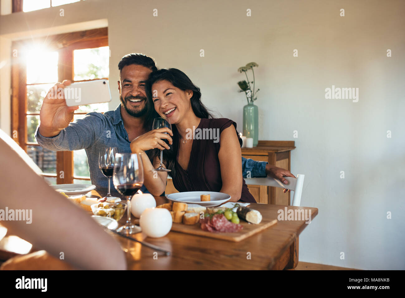 Cheerful couple taking selfie while having dinner with friends at home. Smiling man and woman sitting together at dining table and taking selfie durin Stock Photo