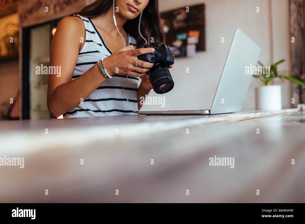 Woman blogger looking at her DSLR camera while working on her laptop computer at home. Woman sitting in front of her laptop with earphones operating a Stock Photo