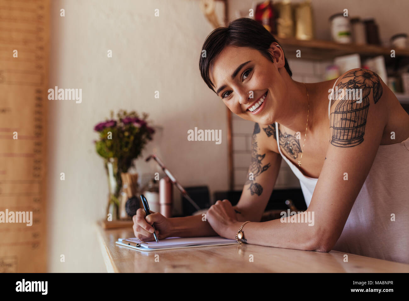 Smiling woman at the counter of her cafe with a pen and pad. Restaurant owner making notes standing at the billing counter. Stock Photo