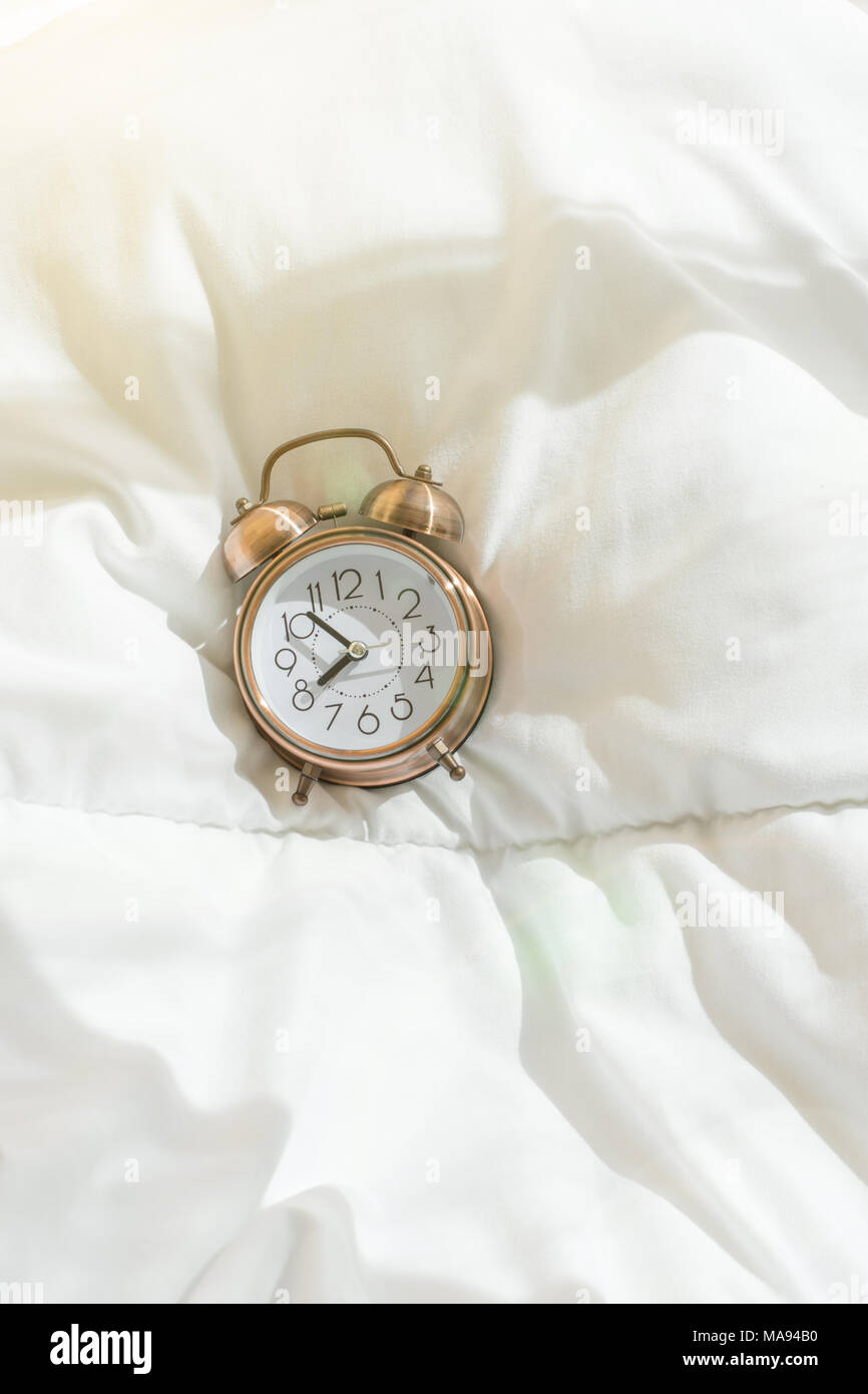 Alarm Clock Showing Eight O'Clock Lying on White Bed Blanket in Bedroom. Bright Morning Sunlight Streaming Through Window. New Day Beginning Waking Up Stock Photo