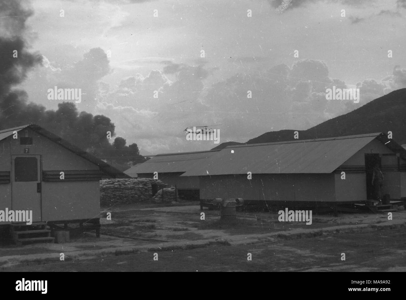 Black and white photograph showing several barracks, and a sandbag bunker, with a soldier walking through a door to the barrack at far right, and a helicopter hovering in the sky in the background, photographed in Vietnam during the Vietnam War (1955-1975), 1968. () Stock Photo