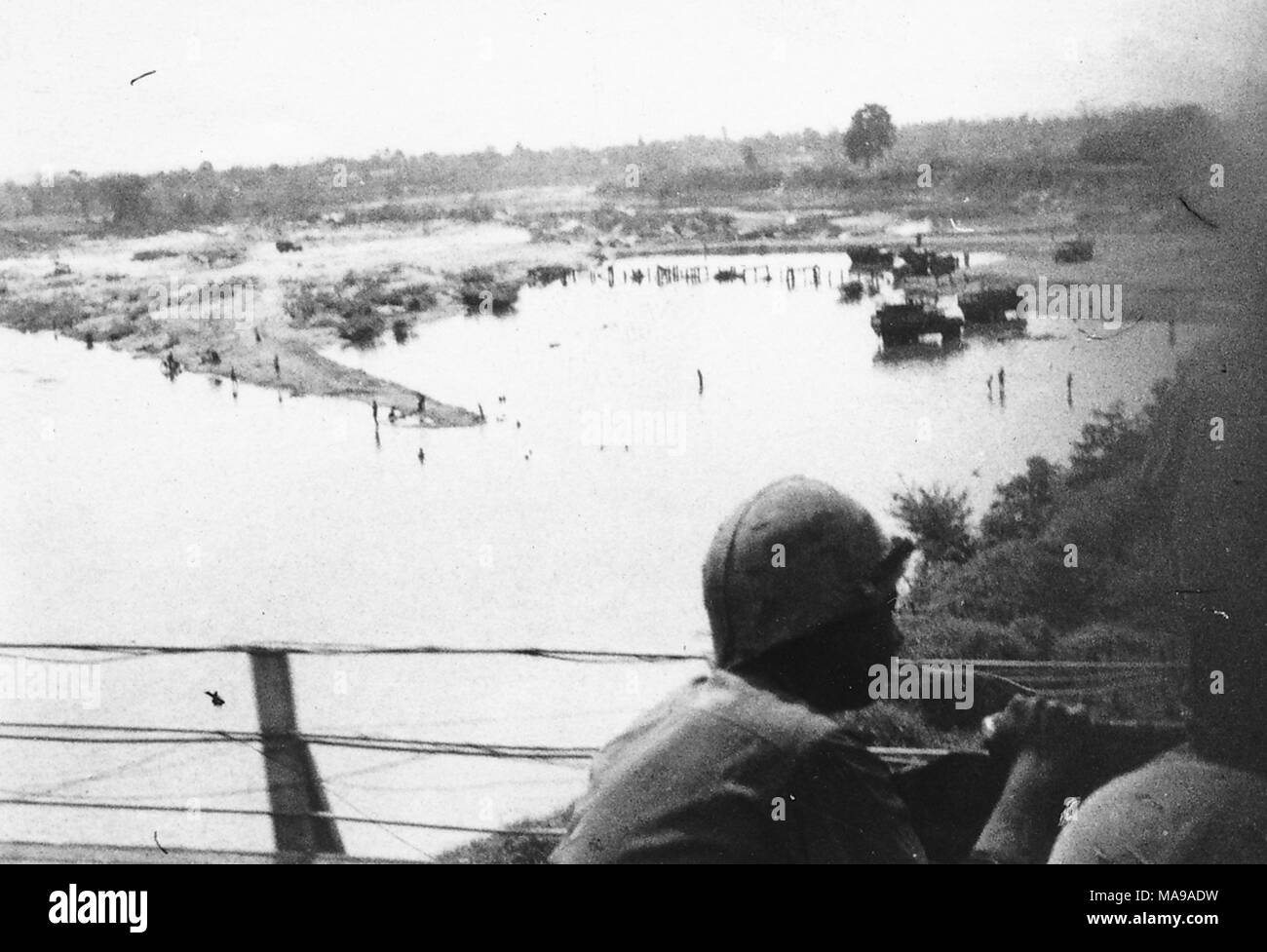 Black and white photograph, showing the back of a helmeted soldier, presumably riding in a truck over a bridge, with a body of water, boats, flat land, and trees in the background, photographed in Vietnam during the Vietnam War (1955-1975), 1971. () Stock Photo