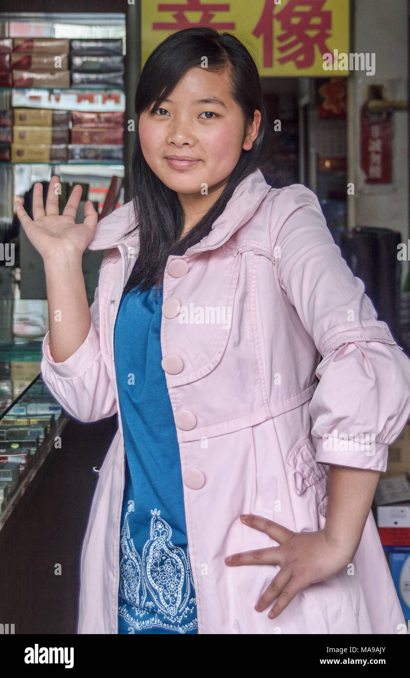 Beijing, China - April 26, 2010: Closeup of young Friendly female store clerk smiling and waving from behind counter. Light pink coat, blue dress. Stock Photo