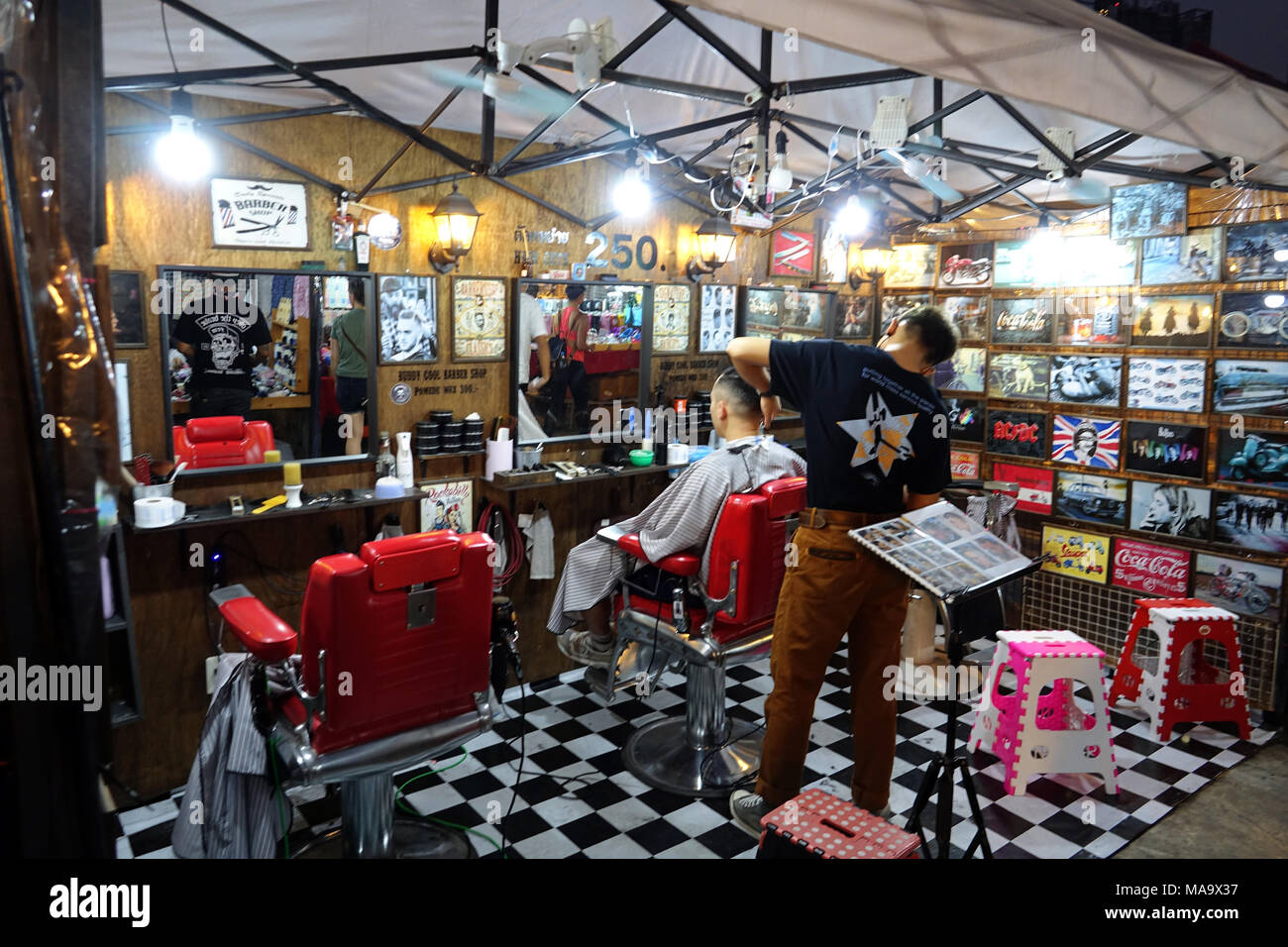 24 February 2018, Thailand, Bangkok: A barber in the Rod Fai Market. The market offers a combination of Thai and street food clothing and bars, making it a very popular destinations for young Thais. Rod Fai Market means translated the Train Market, though nowadays you can't find any trains. The name stayed from the time when the market was directly next to the railway in Chatuchak. This market was dissolved due to construction works. Since 2013 this new market has emerged, the Rod Fai Market Ratchada next to Ratchadaphisek Road. Photo: Alexandra Schuler/dpa Stock Photo