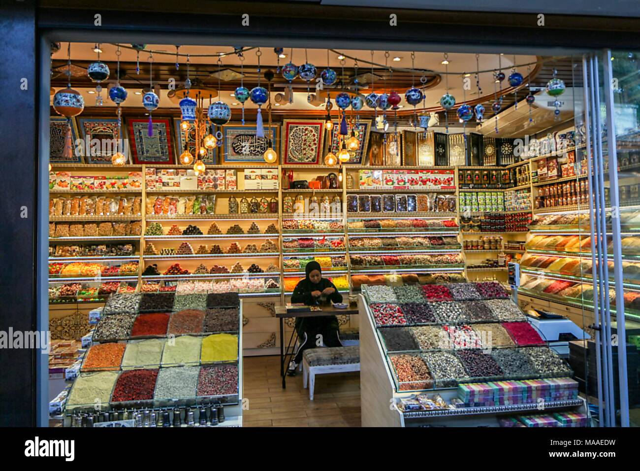 Woman wearing a hijab and track pants, sitting at a table inside a shop filled with colorful souvenirs, ceramics, spices, and other foodstuffs, located in the Arasta Bazaar in the Sultanahmet district of Istanbul, Turkey, November 10, 2017. () Stock Photo