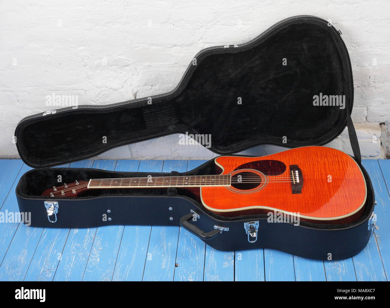 Musical instrument - Orange flame maple, acoustic guitar in hard case on a blue wood, and white brock wall background. Stock Photo
