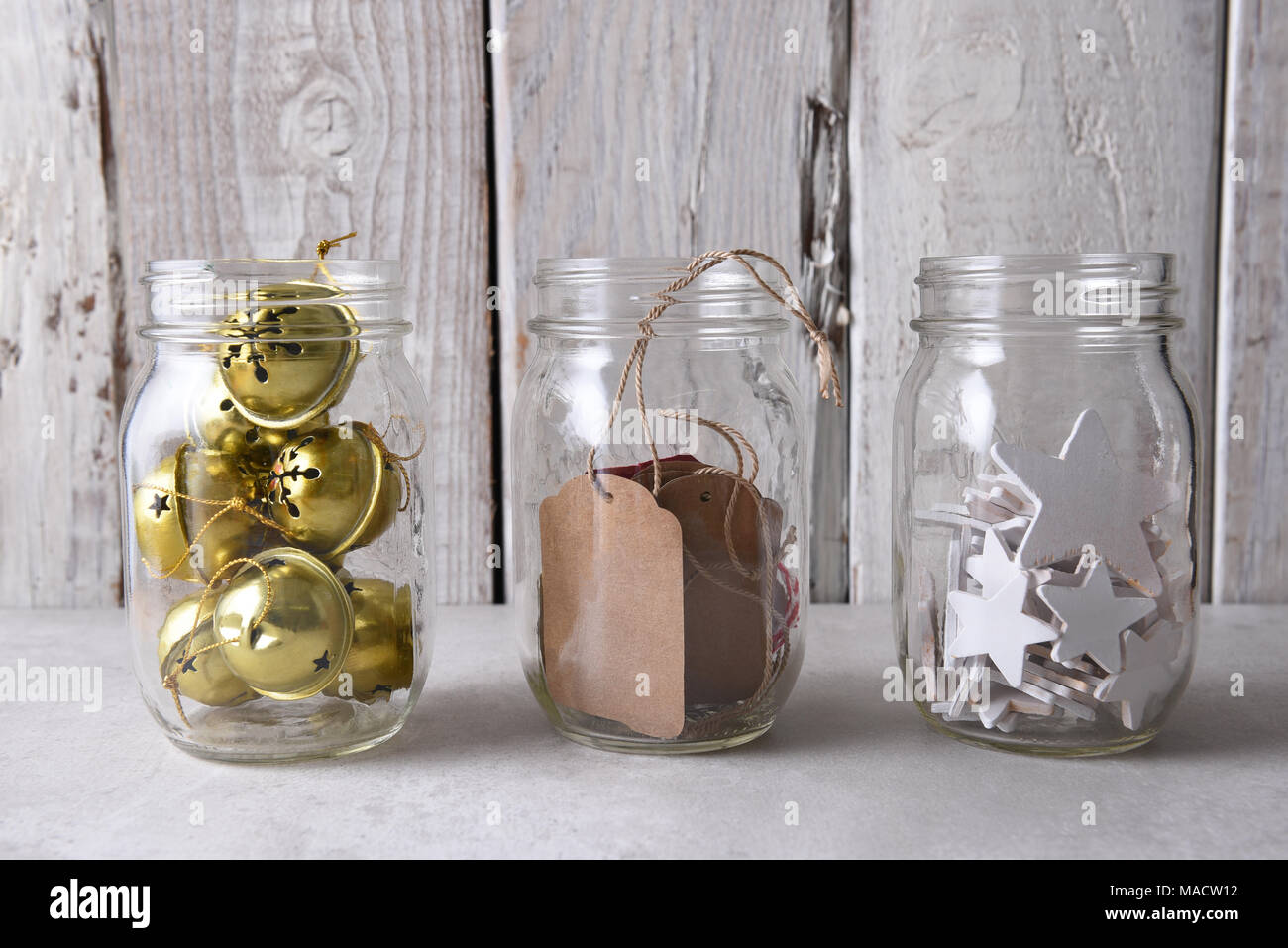 Christmas present wrapping supplies. Three mason jars with gift tags, wood stars, and sleigh bells against a rustic white wood wall. Stock Photo