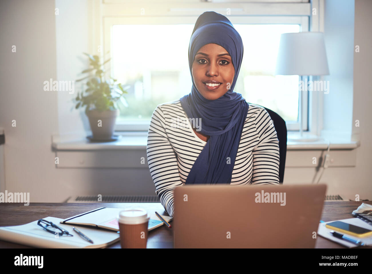 Smiling young Muslim female entrepreneur wearing a hijab sitting in her home office working online with a laptop Stock Photo