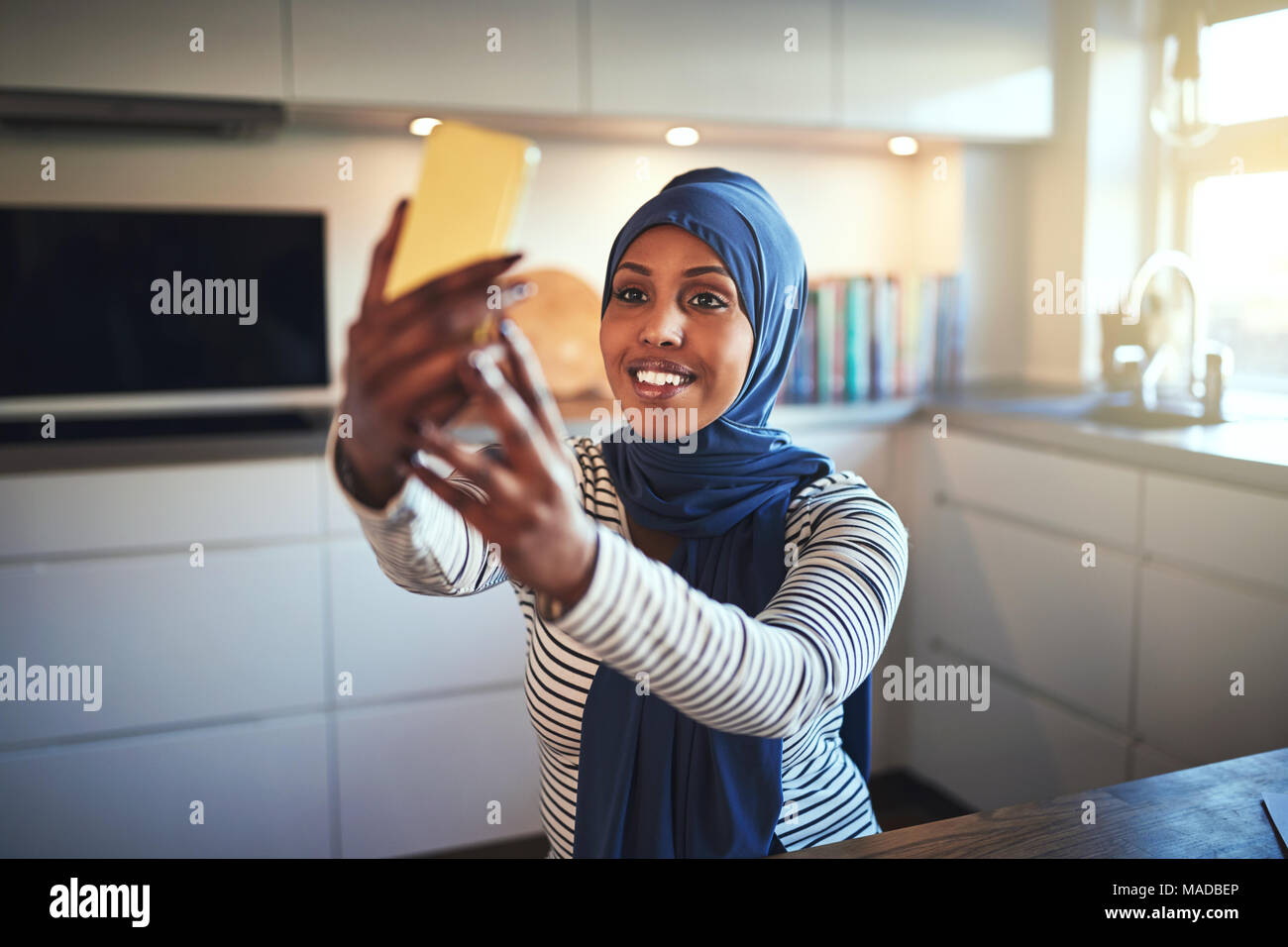 Smiling young Arabic woman wearing a hijab sitting in her kitchen at ...