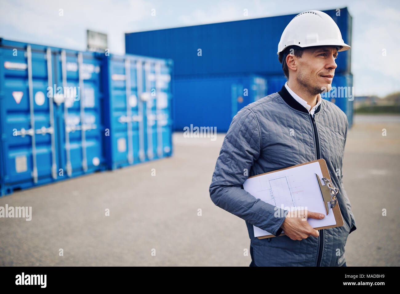 Port manager wearing a hardhat standing alone on a large commercial shipping dock holding a clipboard Stock Photo