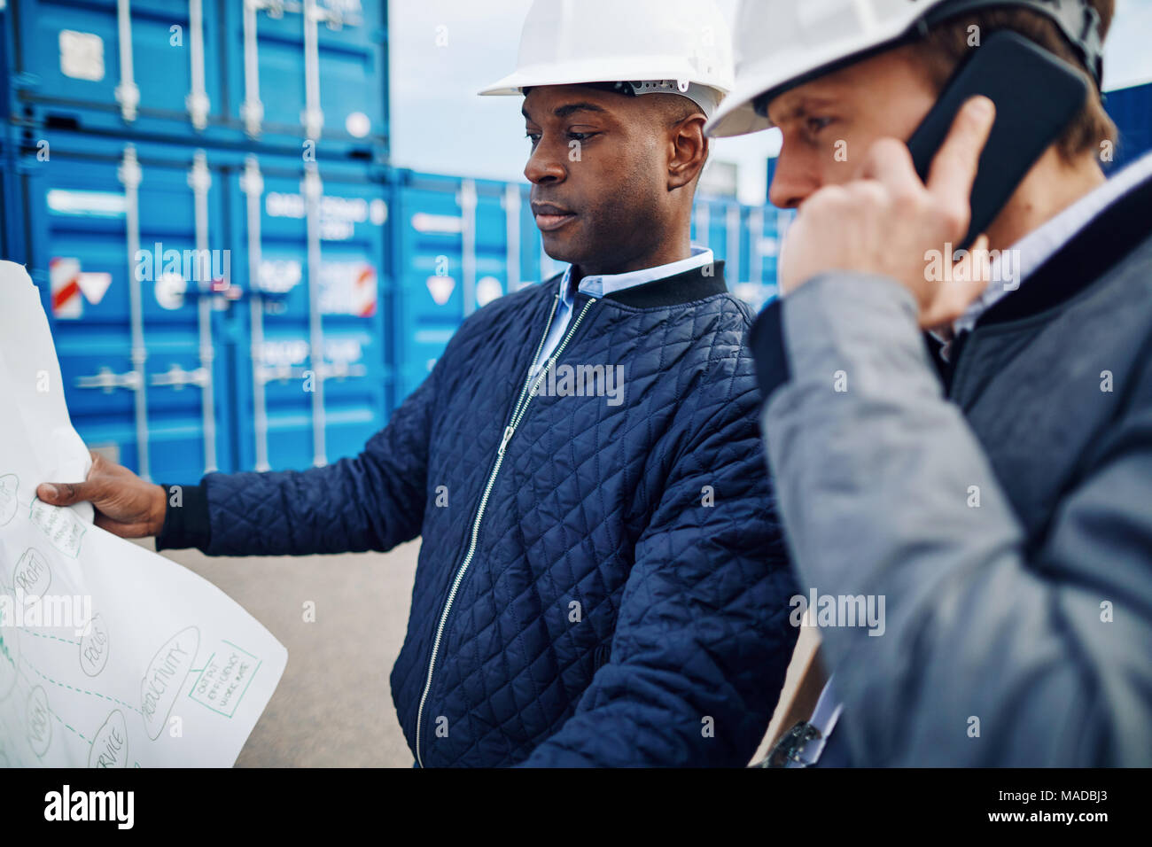 Two engineers holding a building plan and talking on a cellphone while standing by shipping containers in a large commercial shipping yard Stock Photo
