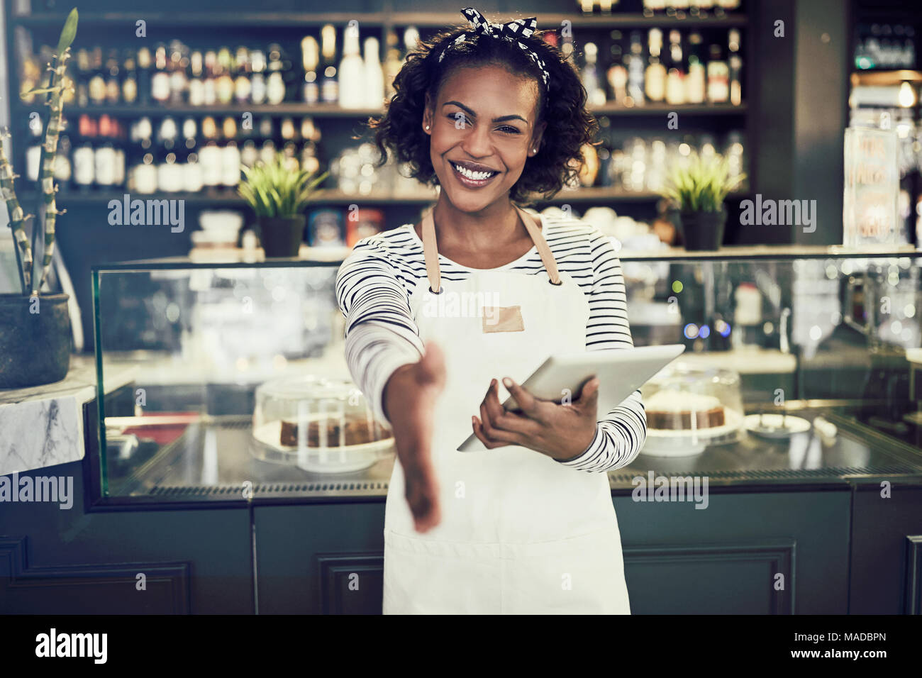 Smiling young African cafe owner extending a handshake while standing in a trendy cafe holding a digital tablet Stock Photo