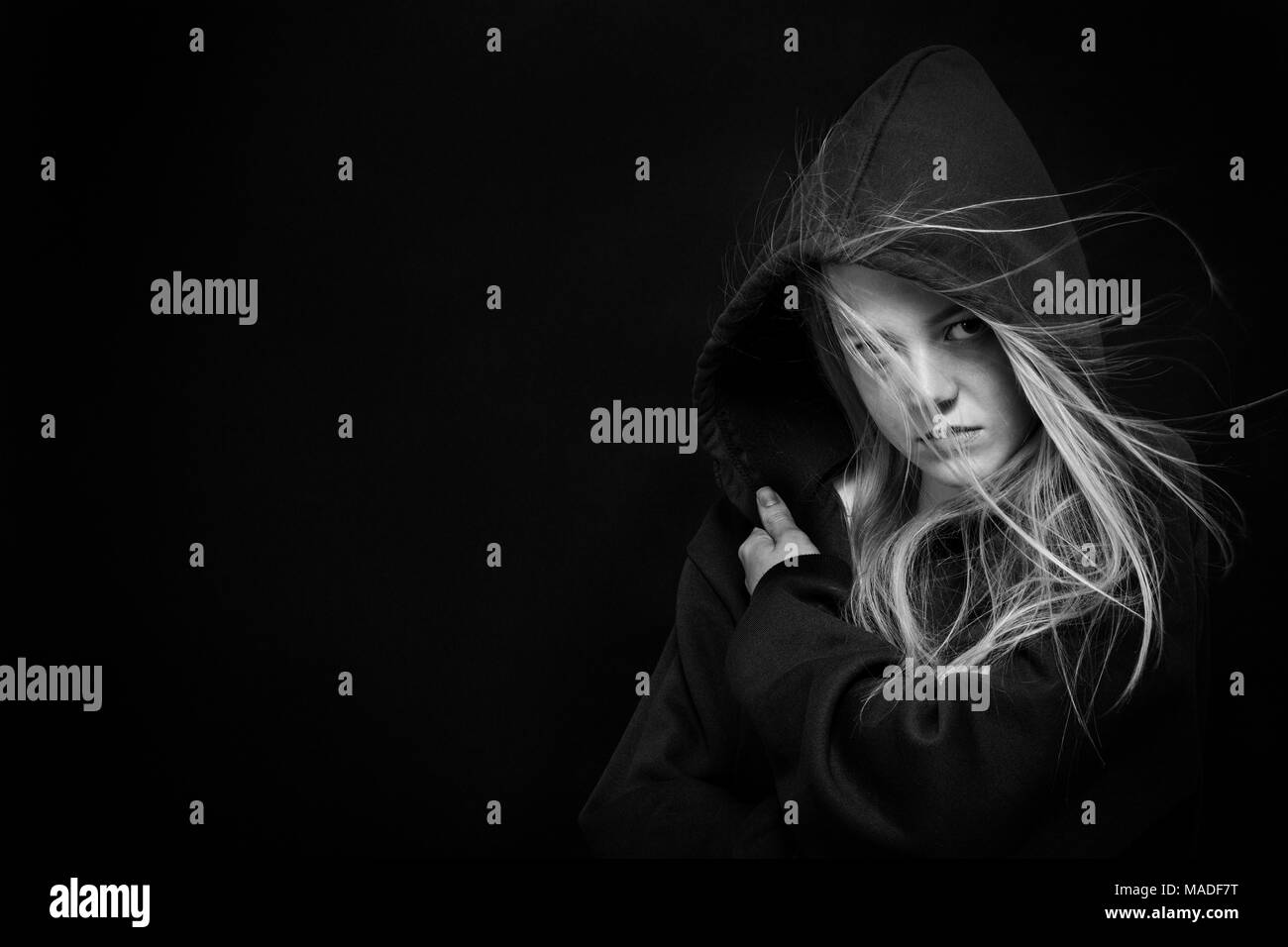 scared blonde girl with flying hairs in hood on black background with copy space looking at camera, monochrome Stock Photo