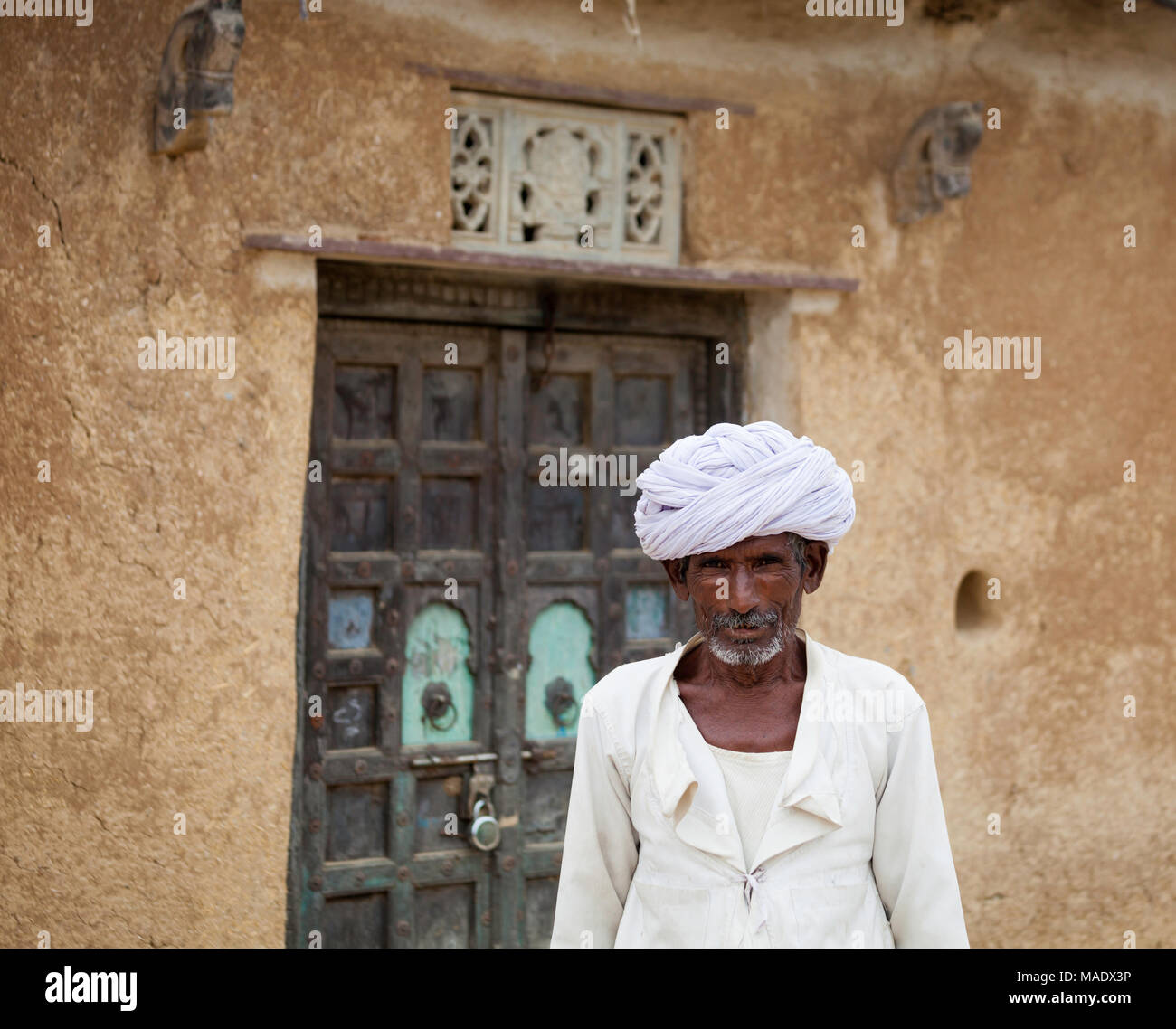 Rajasthani man in white turban and traditional attire, photographed outside a mud-walled building designed in vernacular village style. Stock Photo