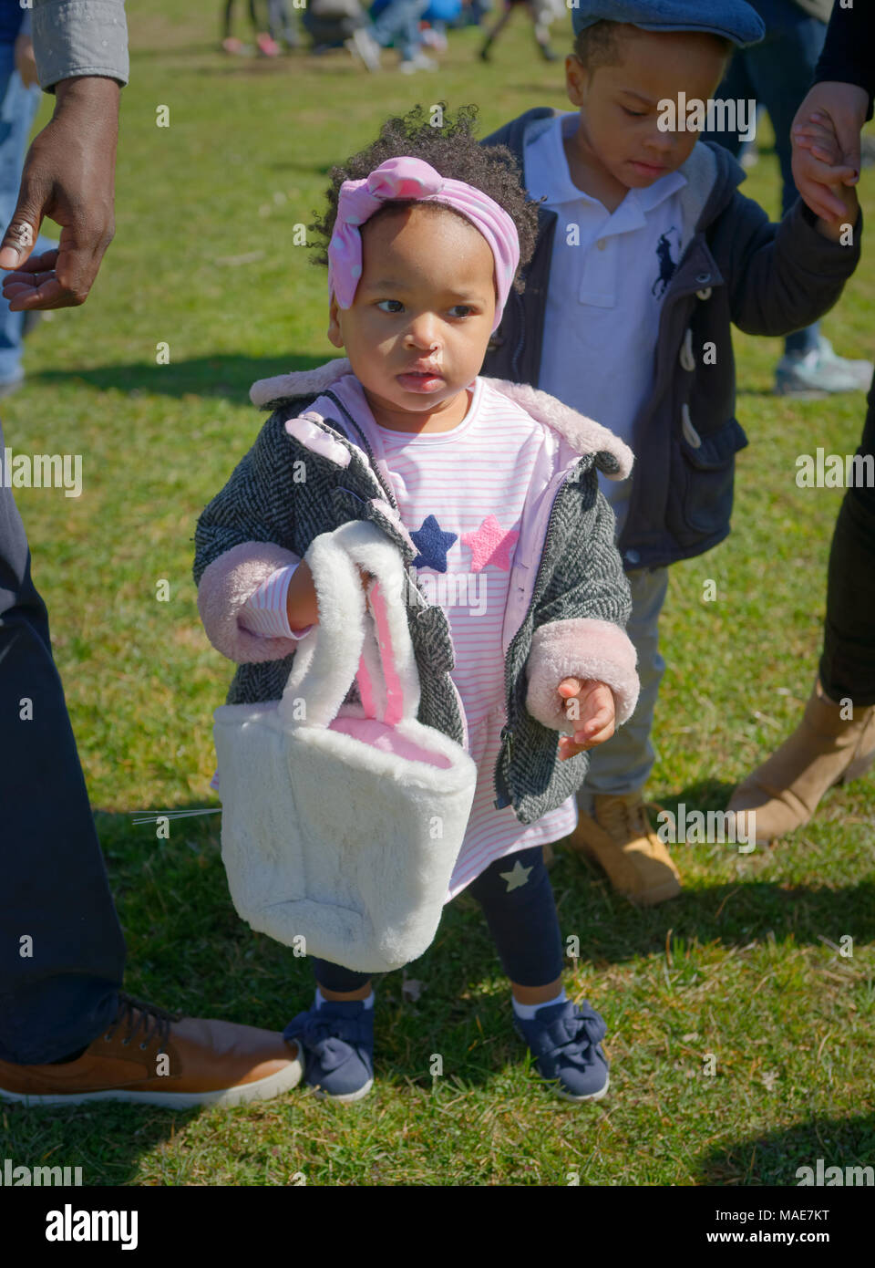 Toddler girl holds her soft white Easter basket as she and her big brother hunt for eggs at the Annual Eggstravaganza, held at Fraser Park and hosted by North and Central Merrick Civic Association (NCMCA). Credit: Ann E Parry/Alamy Live News Stock Photo