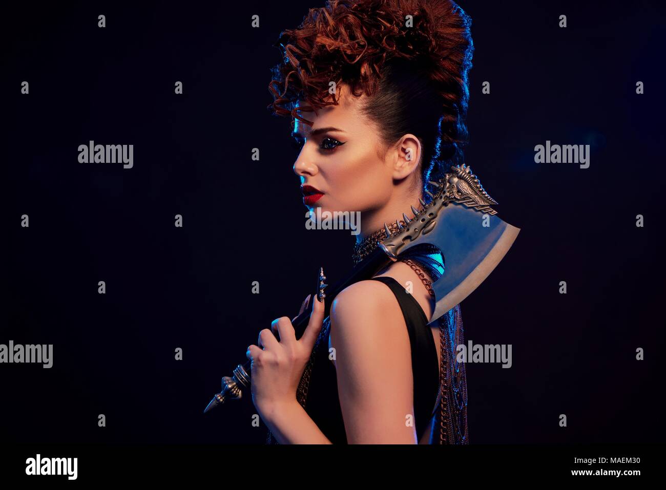 Young beatiful warrior girl holding sharp metallic axe with thorns. Model wears bright original make up and fashionable, stylish hairdress, chains with crosses on her neck. Black studio background. Stock Photo