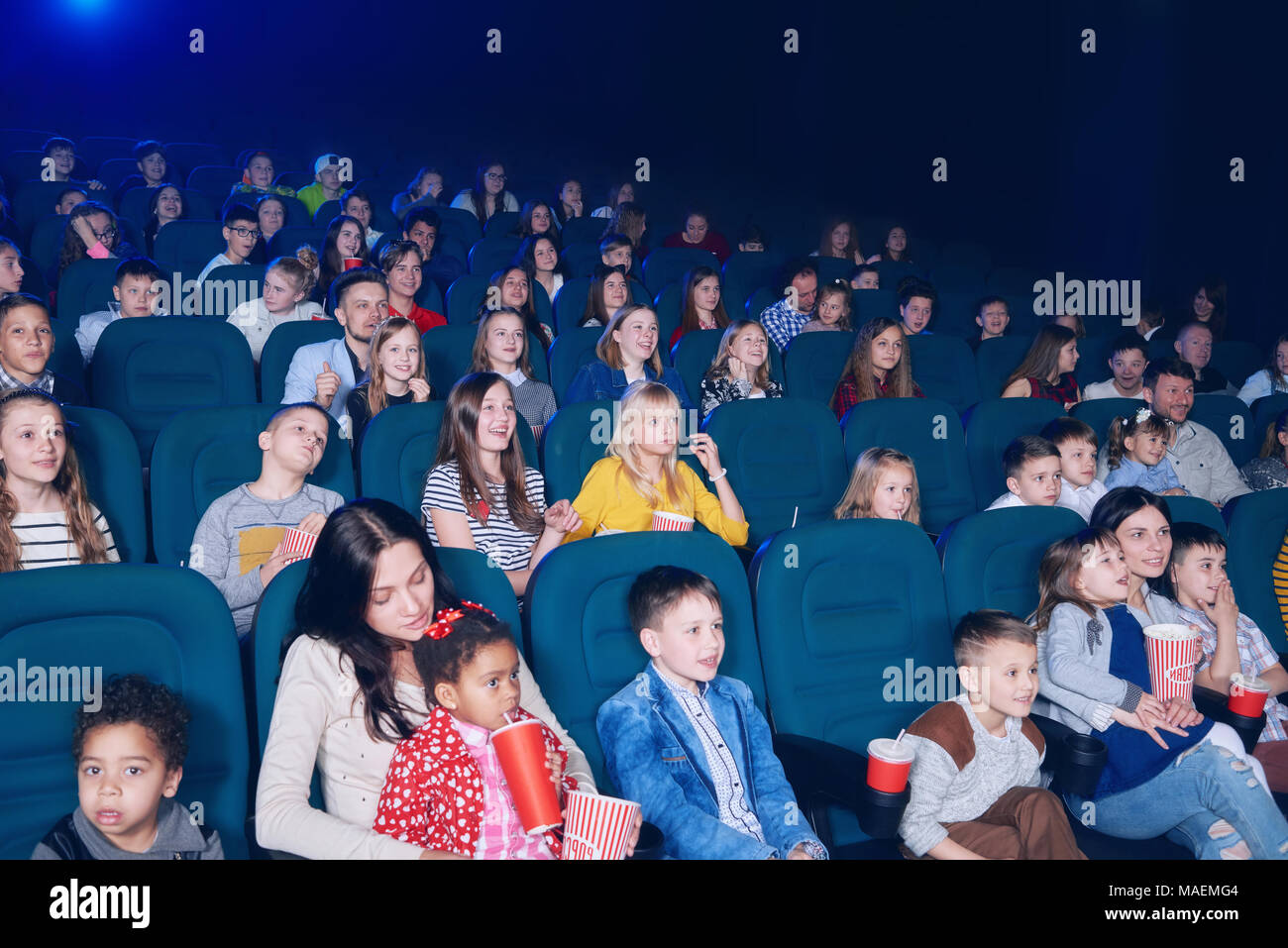 frontview of young exited people watching interesting movie in cinema hall. Boys and girls look very emotional and happy. Models wearing colorful clothes,eating popcorn, drinking fizzy drinks. Stock Photo