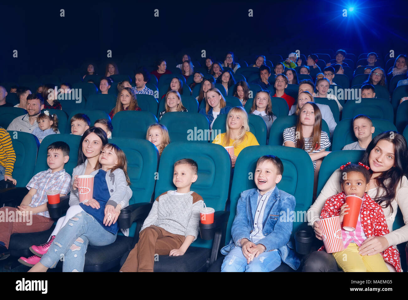 frontview of happy children and their mothers watching interesting, funny movie, sitting on comfortable green places. Boys and girls laughing, smiling, eating popcorn and drinking fizzy drinks. Stock Photo