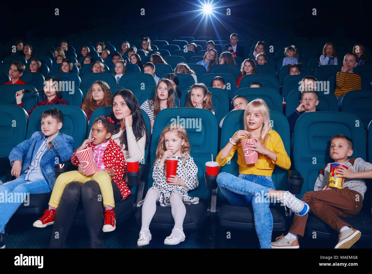 Frontview of emotional people watching film in the new cinema. There are adults and children wearing colorful clothes, keeping fizzy drinks and cola, eating popcorn and smiling. Stock Photo