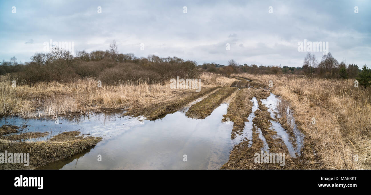 Off-road track with tire imprints and stagnant water in winter landscape. Spring thawing in deserted military area. The muddy bumpy path and blue sky. Stock Photo