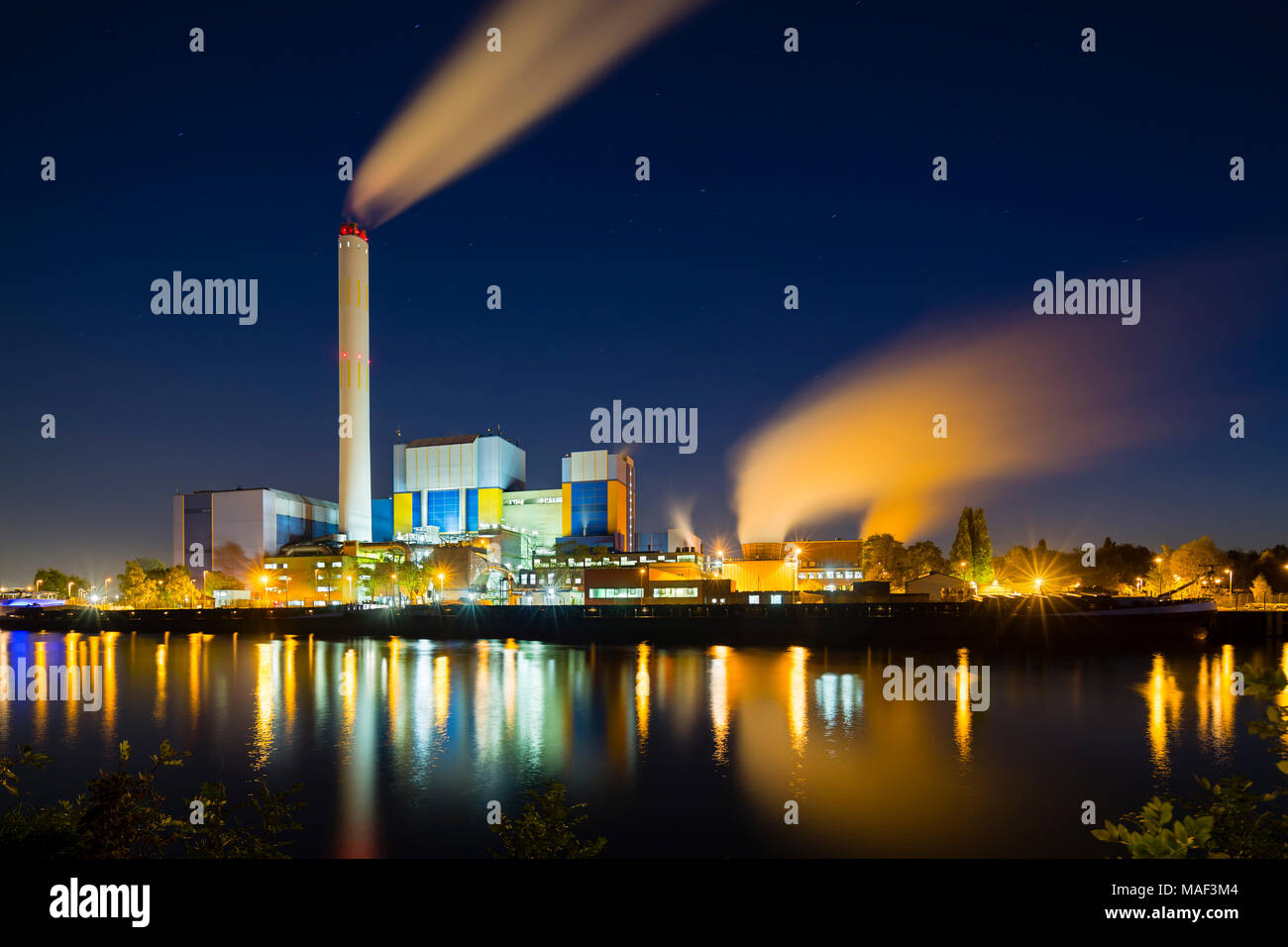 Colorful night shot of an illuminated modern industrial building with steam and deep blue sky behind a canal. Stock Photo