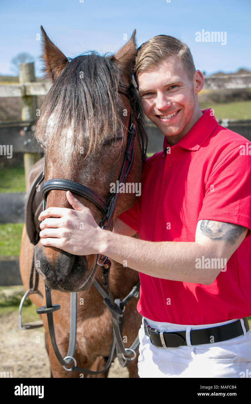 Handsome, smiling horse rider standing next to and petting his horse. Stock Photo