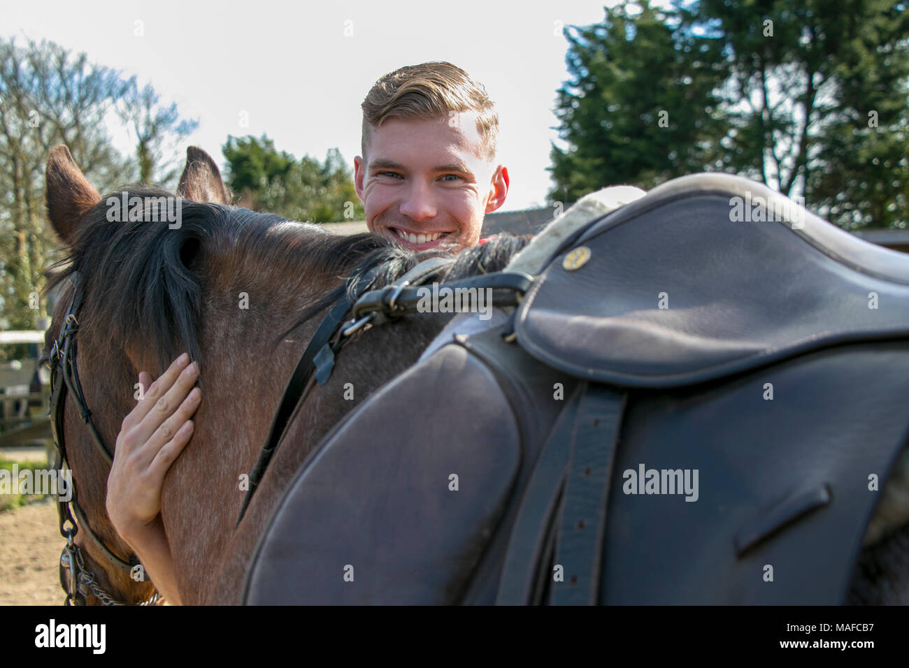 Handsome, smiling horse rider standing next to and petting his horse. Stock Photo