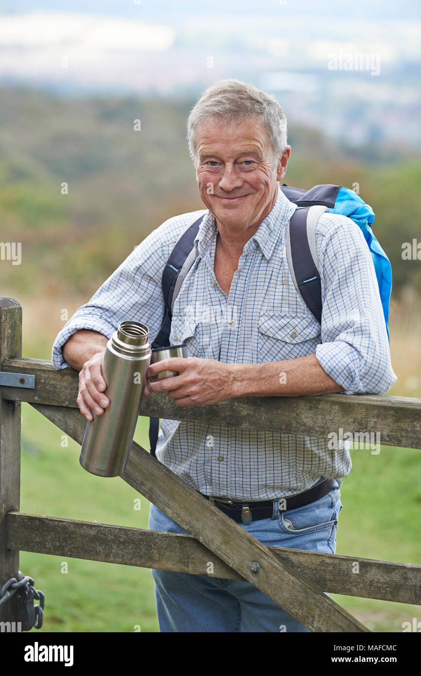 Portrait Of Senior Man On Hike In Countryside Resting By Gate And Having Hot Drink Stock Photo