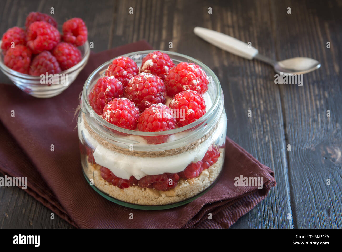 Raspberry cheesecake dessert in glass jar with fresh raspberries and cream cheese on wooden background. Healthy homemade summer berry layered dessert. Stock Photo