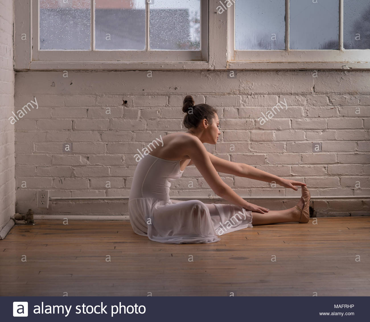 Ballerina Stretching On A Wood Floor With A Brick Wall In The