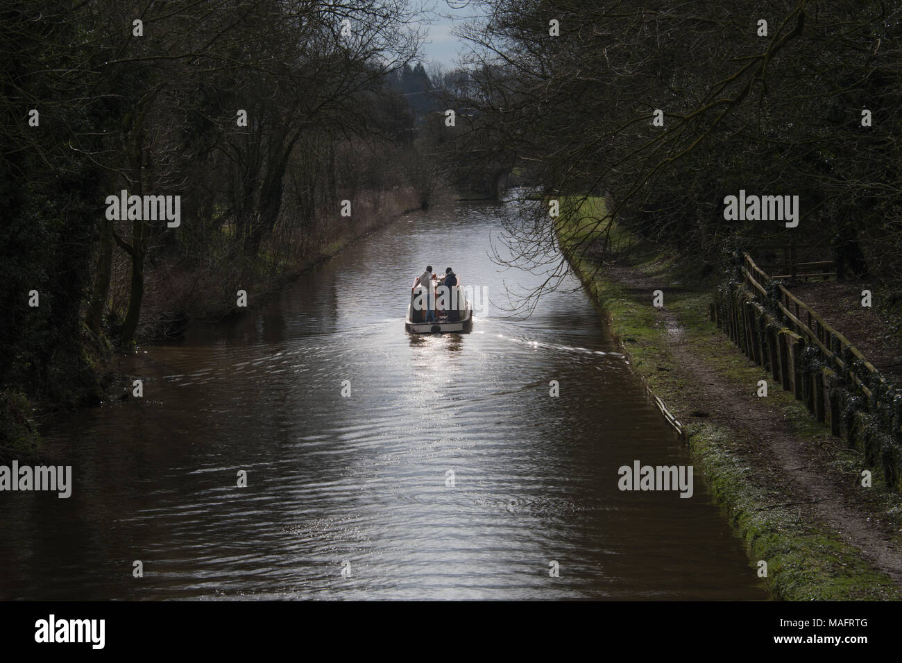 Staffordshire, UK - April 1st 2018 - a scenic view of a  narrowboat on a canal travelling away from the viewer into the sunlight with two young people Stock Photo