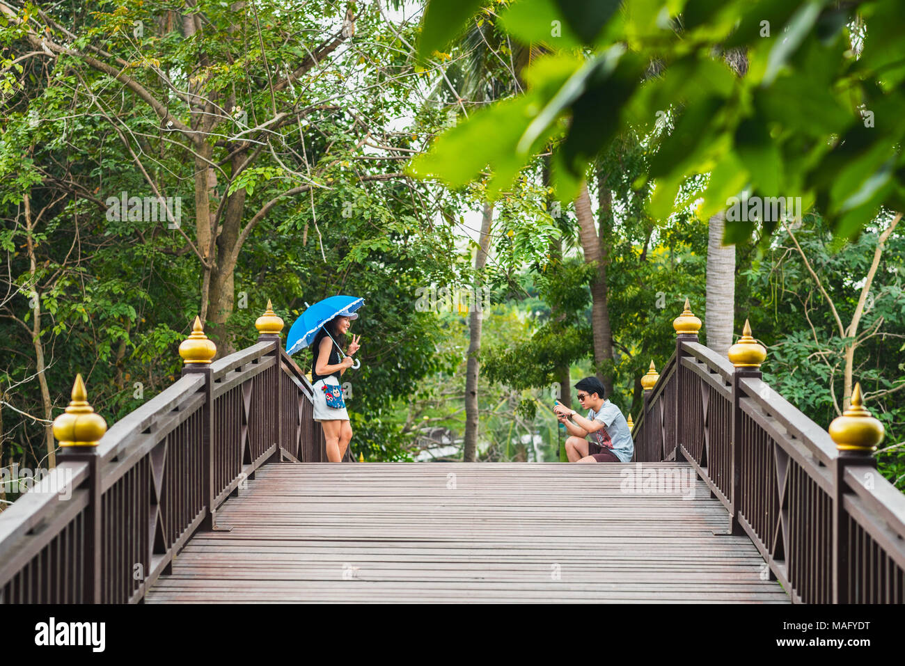 Young Asian man photographs a smiling young Asian woman with blue umbrella on wooden bridge in park in Bang Krachao (Bang Kachao), Bangkok, Thailand. Stock Photo
