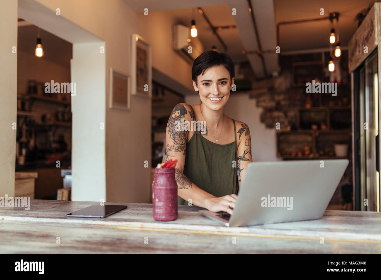 Woman entrepreneur working on laptop computer at home with a smoothie on the table. Woman enjoying a glass of smoothie while working at home. Stock Photo