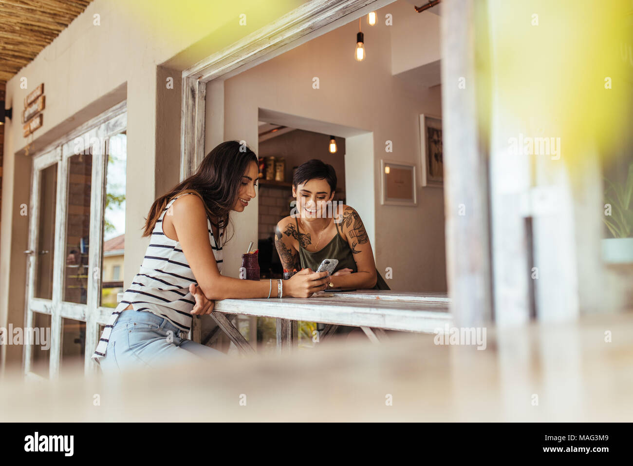 Women looking at mobile phone sitting at a restaurant table. Friends having smoothie and dessert sitting at a restaurant. Stock Photo