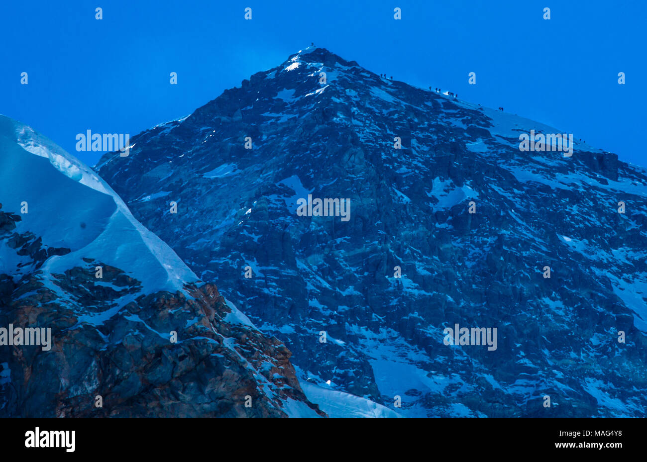 Snow can be seen blowing past the tiny backlit figures of climbers from both Nepal and Tibet reach the summit of Mount Everest, Nepal. Stock Photo