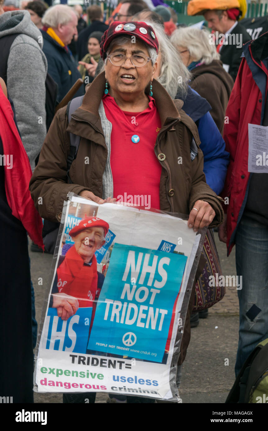Aldermaston, UK. 1st Apr, 2018. A singer from Welsh socialist choir Côr Cochion holds a poster 'NHS Not Trident' and sings as CND celebrates the 60th anniversary of the first Aldermaston march which mobilised thousands against the Bomb and shaped radical protest for generations. Their protest outside the Atomic Weapons Establishment included a giant, iconic peace symbol, speeches, including by some of those on the original march, singing and drumming and celebrated the UN treaty banning nuclear weapons. Credit: Peter Marshall/Alamy Live News Stock Photo
