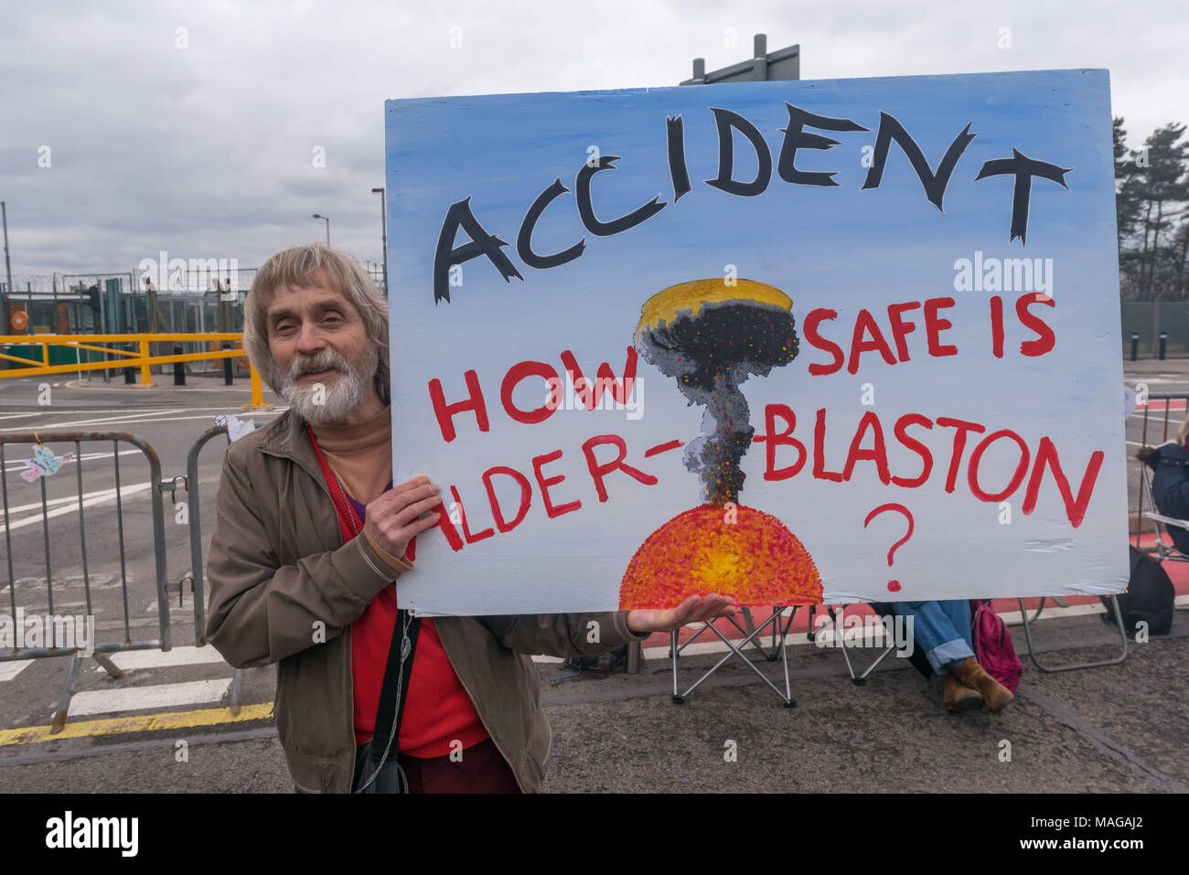 Aldermaston, UK. 1st Apr, 2018. A campaigner holds up a large poster with the message 'Accident - How safe is Alder-Blaston' painted specially for the CND celebration of the 60th anniversary of the first Aldermaston march which mobilised thousands against the Bomb and shaped radical protest for generations. Their protest outside the Atomic Weapons Establishment included a giant, iconic peace symbol, speeches, including by some of those on the original march, singing and drumming and celebrated the UN treaty banning nuclear weapons. Credit: Peter Marshall/Alamy Live News Stock Photo
