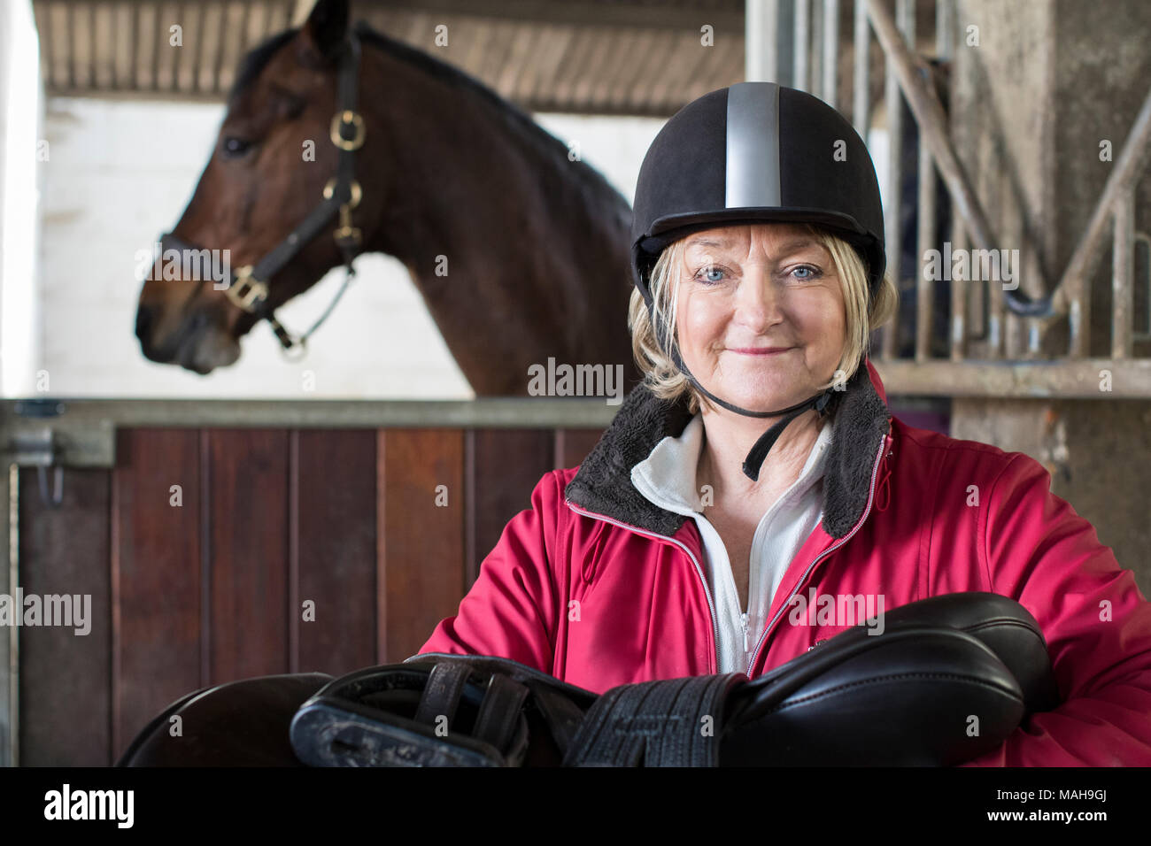 Portrait Of Mature Female Owner Holding Saddle In Stable With Horse Stock Photo