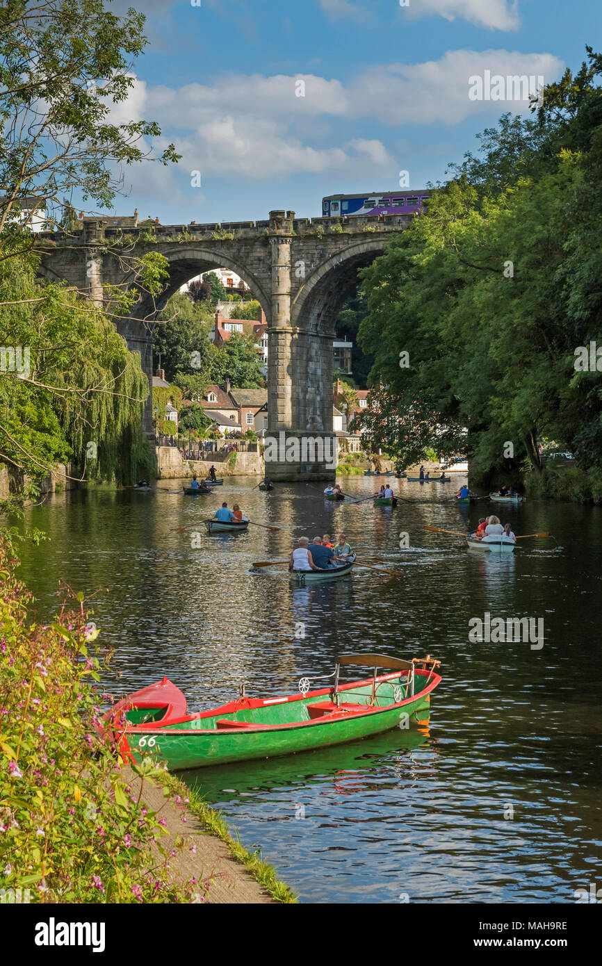 People relaxing & boating in rowing boats on River Nidd under blue summer sky, as train passes over bridge in scenic sunny  Knaresborough, England, UK Stock Photo