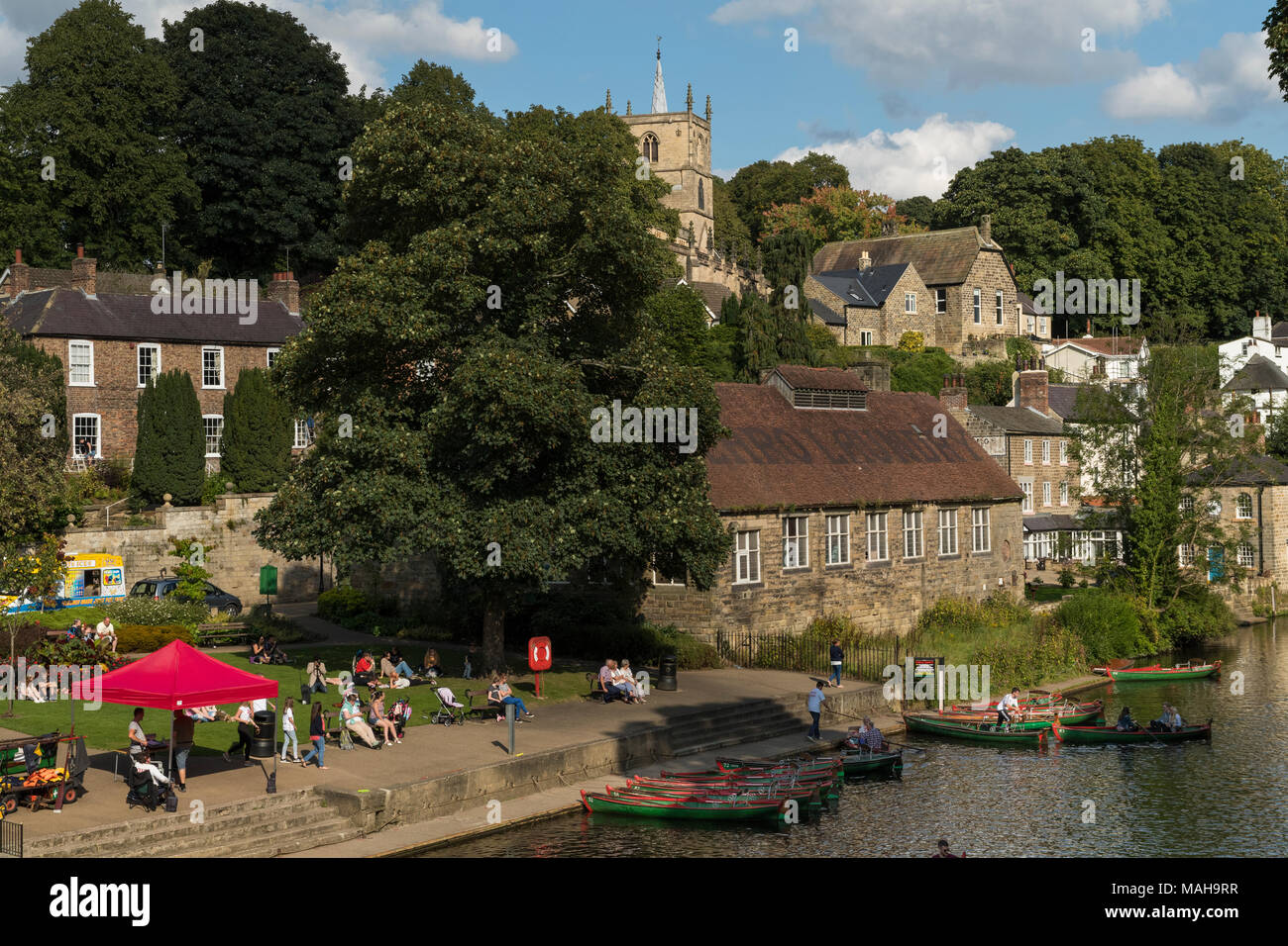 People relaxing by riverside & boating in rowing boats on River Nidd under blue sky - scenic sunny summer view by bridge, Knaresborough, England, UK. Stock Photo