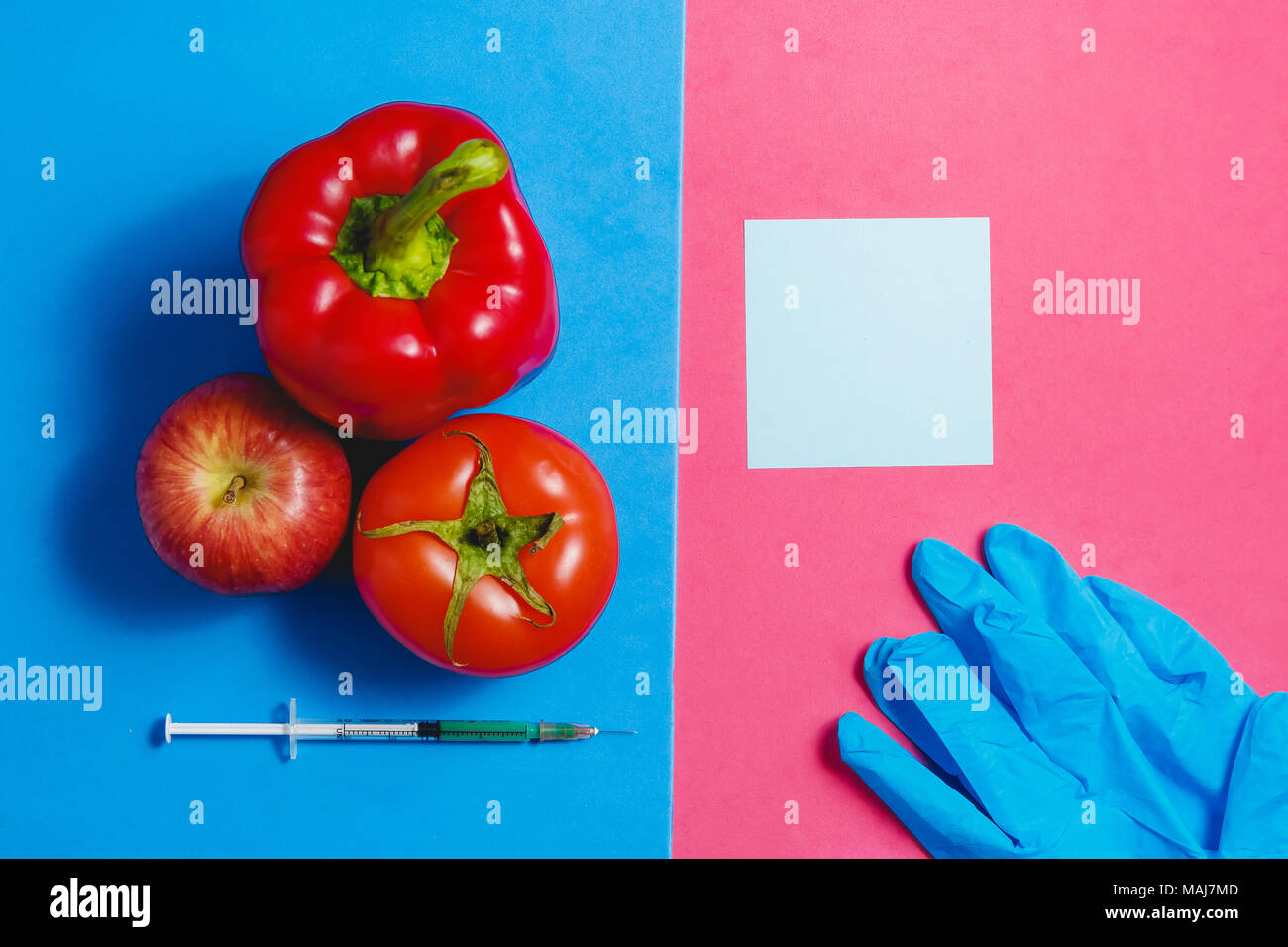 Note, Green Liquid in Syringe, Red Tomato, Apple, Pepper, Blue Gloves. Genetically Modified Food Concept on Pink Blue. Stock Photo