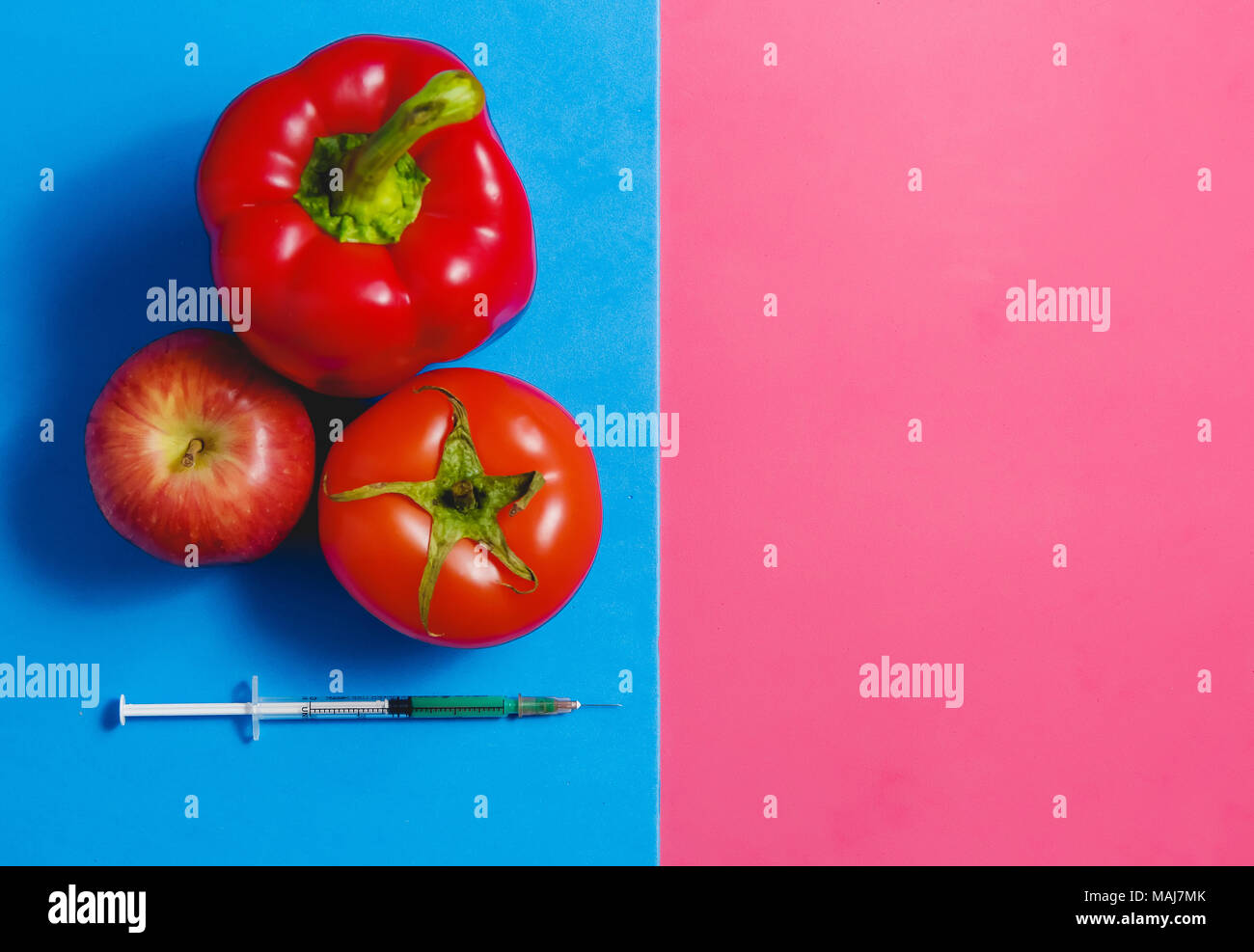 Green Liquid in Syringe, Red Tomato, Apple, Pepper. Genetically Modified Food Concept on Pink Blue. Stock Photo