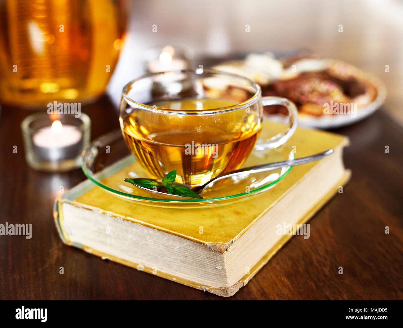 Teatime scene with glass tea pot and cup of tea, old book and hot steam. Idyllic tea time or relaxation scene with cookies and candles. Stock Photo