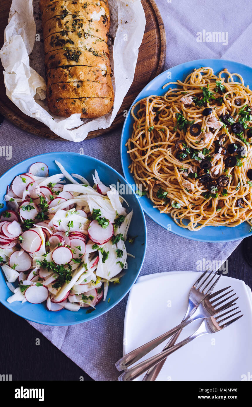 Traditional Italian dinner - Spaghetti Alla Puttanesca pasta with black olives, tuna, anchovies, capers and parsley, vegetable salad and baked ciabatt Stock Photo