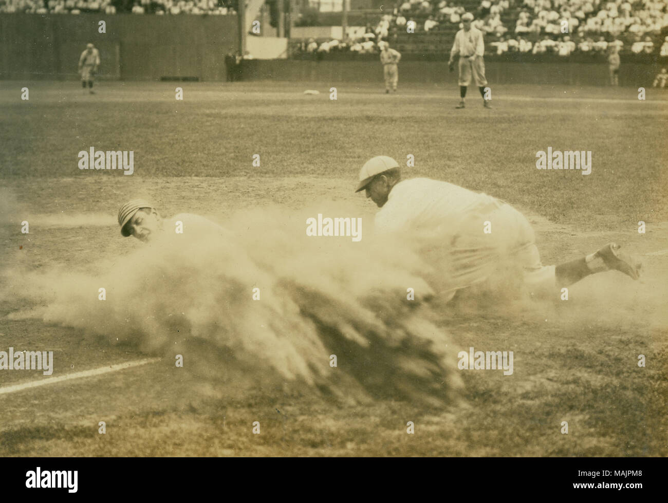 A man is sliding into first base with the first basman there trying to tag him out. The dirt is flying everywhere. Can see the outfielders. Title: Baseball player sliding into third base at a St. Louis Terriers (Federal League) game.  . between 1914 and 1915. Russell Froelich Stock Photo