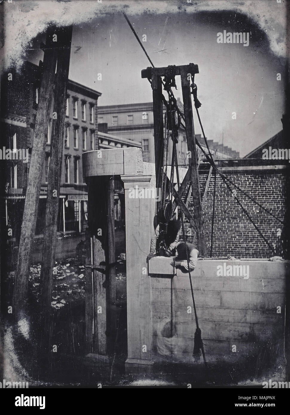 View of the northeast corner of Fourth and Olive Streets of the construction of a marble building. Two workmen use a hoist to fit a block into the wall. Commercial buildings can be seen in the background, as well as stone rubble in the street. Title: Construction of Marble Building, Northeast Corner of Fourth and Olive Streets, Workmen with Hoist fitting Block into Wall.  . 1850. Thomas M. Easterly Stock Photo