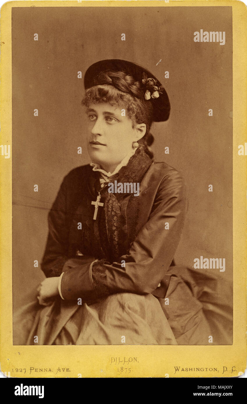 Cabinet card studio photograph of Anna J. Barnes; she is leaning on draped photographer's prop. Anna J. Barnes is wearing a flat hat with hanging ornamentation, has a row of braided hair below the hat, and curled bangs. Her dress appears to be heavy satin with lace trim around the cuff, down the front, and at the collar. There is a white ruffled inset at her throat, and she wars a large cross pendant and bobble earrings. It is a three-quarter pose looking to the viewer's left. Title: Anna J. Barnes.  . 1870. Dillon, Washington, DC Stock Photo